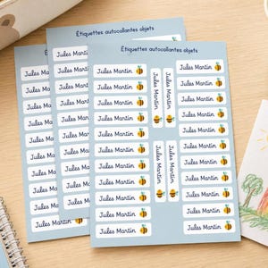 Peut inclure: Trois feuilles d'étiquettes autocollantes bleu clair pour objets, comportant chacune plusieurs vignettes rectangulaires blanches imprimées avec le nom Jules Martin et une petite illustration d'abeille jaune et noire, avec le texte Étiquettes autocollantes objets.