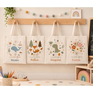 Peut inclure: Quatre sacs fourre-tout en toile blanc cassé suspendus à une étagère en bois. Chaque sac présente une illustration unique : une baleine bleue, un renard en forêt, un dinosaure vert et un bouquet floral. Sous chaque motif, le texte mes petites affaires.