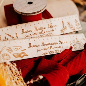 Peut inclure: Deux règles en bois avec texte gravé et éléments décoratifs. L'une dit "Merci Maître Alban pour cette belle année! 2022-2023". L'autre dit "Merci Maîtresse Anne pour cette belle année! Julie 2022-2023". Une bobine de ruban rouge est en arrière-plan.