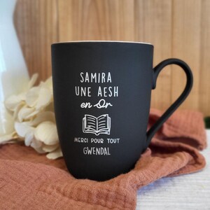 Op de afbeelding: Een zwarte keramische mok met een witte binnenkant en een gebogen handvat. De mok heeft witte tekst met de tekst "SAMIRA UNE AESH en Or", een open boekillustratie en "MERCI POUR TOUT GWENDAL". De mok ligt op een perzikkleurige doek.