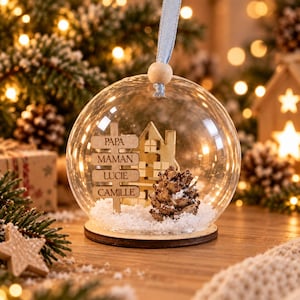 Peut inclure: Une boule de No&euml;l en verre transparent contenant un panneau en bois grav&eacute; avec les noms PAPA, MAMAN, LUCIE et CAMILLE. &Agrave; l'int&eacute;rieur, une silhouette de maison en bois et une pomme de pin reposent sur de la neige artificielle blanche.