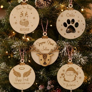 Peut inclure: Cinq décorations de Noël circulaires en bois avec des attaches en ficelle rouge et blanche. Les motifs incluent un renne avec le texte Mon 1er Noël ARTHUR, une patte avec Oslo, une tête de renne avec Joyeux Noël Jules, des ailes d'ange avec Papy Fred et un Père Noël avec Victoire.