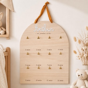 Op de afbeelding: Houten baby mijlpaalbord met de naam "Arthur" in witte letters. Het bord heeft gouden clips voor foto's, gemarkeerd met "1 mois" tot "12 mois". Een mosterdkleurig lint wordt gebruikt om op te hangen.
