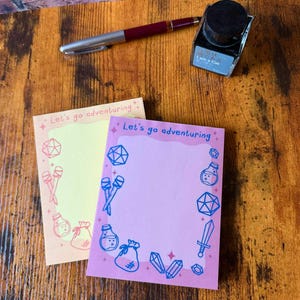 May include: Two notepads with the text "Let's go adventuring" and fantasy-themed illustrations. One notepad is light yellow, the other is pink. A burgundy pen and a bottle of black ink are also on the wooden surface.