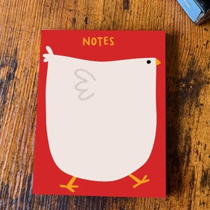 May include: A red notepad with the word "NOTES" at the top. The notepad features a whimsical illustration of a white chicken with a yellow beak and feet. The chicken design is centered on the notepad.