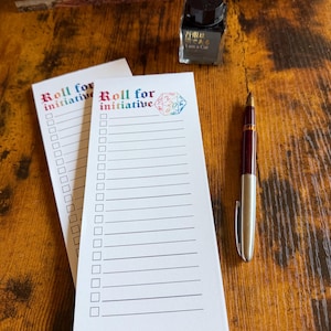 May include: Two white notepads with the text "Roll for initiative" in colorful lettering. One notepad has a D20 die graphic. A burgundy and silver pen and a bottle of black ink are also on the wooden surface.