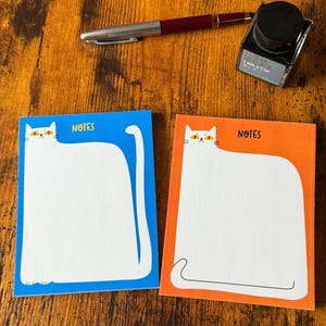 May include: Two cat-shaped notepads, one blue and one orange, with the word "NOTES" printed on them. A pen and ink bottle are on a wooden surface. The notepads have a white cat design with yellow eyes.