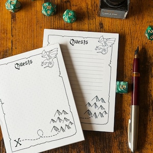 May include: Two white "Quests" notepads with a dragon illustration, mountain range, and dotted path design. A burgundy fountain pen, green dice, and inkwell are on a wooden surface. The notepad has a dotted grid.