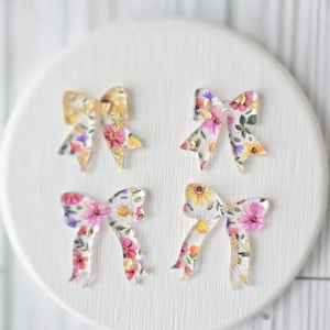 May include: Four clear resin bows with a floral print. The bows are in two different sizes and have a gold glitter accent.