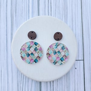 May include: A pair of round earrings with a colorful geometric pattern in shades of pink, teal, and gold. The earrings are attached to small, dark brown wooden studs. The earrings are displayed on a white, round wooden base.