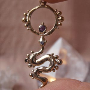 Puede incluir: Un colgante dorado con forma de serpiente estilizada. El colgante presenta una parte superior circular con una gema púrpura y un cuerpo sinuoso con pequeños adornos redondos. El cuerpo de la serpiente se curva con gracia, y el colgante es sostenido por un dedo.