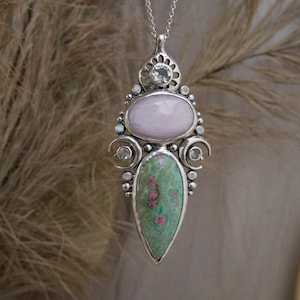 May include: A silver pendant necklace with a green and pink gemstone, a pink oval gemstone, and clear gemstones. The pendant is in the shape of a teardrop.
