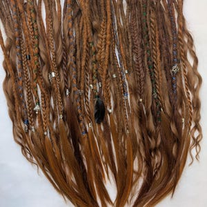 May include: A collection of brown and auburn dreadlocks, some with braided sections and decorative beads. A few dreads have blue and green accents, and a black feather is included. The dreadlocks are arranged on a white surface.