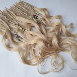 May include: Blonde dreadlocks with wavy ends, decorated with beads and charms. The dreadlocks are a light beige colour, with some wrapped in green thread. The charms include a gold coin, a Hamsa hand, and other small pendants.