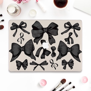 May include: A laptop computer with a black and white design of various sized bows on the lid. The laptop is on a white surface with makeup brushes and candles.