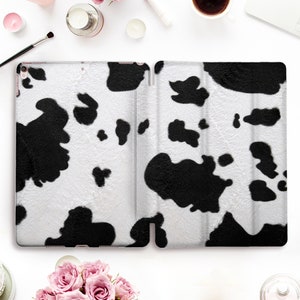 May include: A white and black cow print tablet case, open and displayed. The case has a soft, textured appearance with large black spots on a white background. The case is designed to protect a tablet.