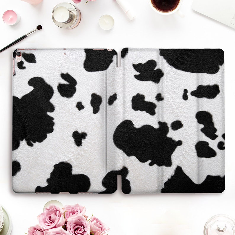 May include: A white and black cow print tablet case, open and displayed. The case has a soft, textured appearance with large black spots on a white background. The case is designed to protect a tablet.