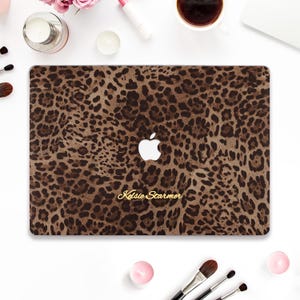 Puede incluir: Una funda para portátil con un diseño de estampado de leopardo marrón. El logotipo de Apple es blanco. El nombre "Kelsie Starmer" está impreso en oro. Pinceles de maquillaje, una taza de café y productos de belleza rodean el portátil.