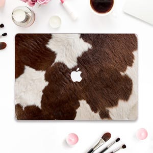 Può includere: Una cover per laptop con un motivo a pelle di mucca marrone e bianca. La cover presenta un logo Apple bianco al centro. Il design ricorda la pelliccia naturale degli animali, offrendo un'estetica rustica e unica. Questa cover protegge il laptop.