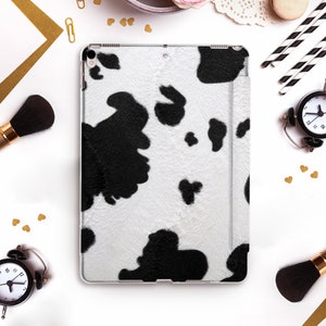 May include: A white and black cow print iPad case. The case has a clear border and a textured pattern resembling cowhide. The case is surrounded by makeup brushes, a clock, and other decorative items.