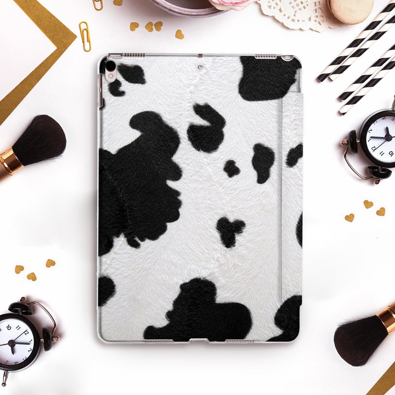May include: A white and black cow print iPad case. The case has a clear border and a textured pattern resembling cowhide. The case is surrounded by makeup brushes, a clock, and other decorative items.