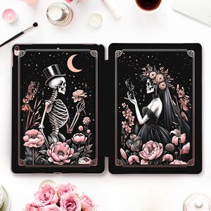 May include: Black tablet case with a skeleton in a top hat and a skeleton bride, surrounded by pink flowers and a crescent moon. The design has a dark, gothic aesthetic with a decorative gold border.