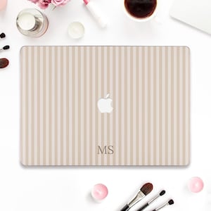 Peut inclure: Coque d'ordinateur portable à rayures verticales beiges et blanches, avec le logo Apple et les initiales "MS". L'image comprend des pinceaux de maquillage et une tasse de café, suggérant un thème de beauté ou de style de vie.
