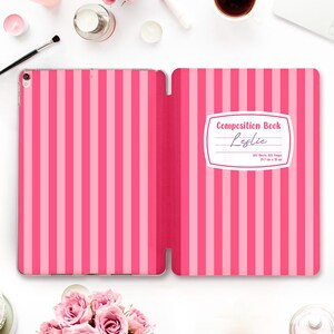 Può includere: Una custodia per tablet con un motivo a righe verticali rosa e rosa chiaro. Il lato destro presenta un'etichetta bianca con il testo Composition Book, il nome Leslie e le specifiche 100 Sheets 200 Pages 24.7 cm x 19 cm.
