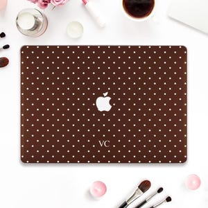 May include: A brown laptop case with a white polka dot pattern. The case features a white Apple logo and the initials "VC" at the bottom. The case is surrounded by makeup and beauty products.