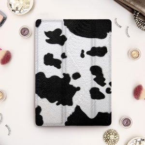 May include: A tablet case with a black and white cowhide pattern. The case is rectangular and features a repeating pattern of black irregular shapes on a white background. The case is surrounded by makeup and accessories.
