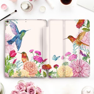 Op de afbeelding: Een witte iPad-hoes met een bloemenontwerp met kolibries en vlinders. De hoes heeft een roze en witte achtergrond met aquarelbloemen en groene bladeren.