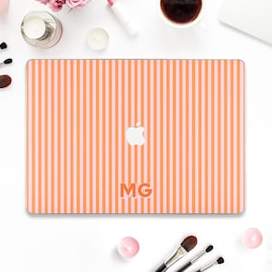 May include: A laptop case with a vertical striped pattern in orange and light pink. The Apple logo is visible, along with the initials "MG" in orange. The case is surrounded by makeup and beauty products.