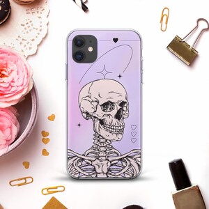 May include: A clear phone case with a purple and pink gradient background. The case features a detailed black and white illustration of a human skull and rib cage. Decorative hearts and stars are also included in the design.