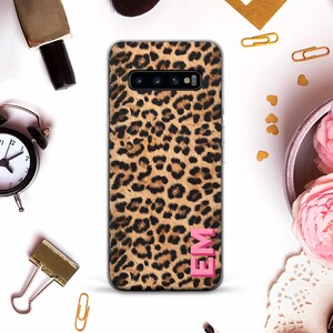 以下が含まれることがあります： ヒョウ柄のデザインのスマートフォンケース。ケースは茶色と黒の斑点模様で、右下にピンク色のイニシャル「EM」が付いています。ケースの周りには、黒い目覚まし時計や金色のペーパークリップなどのアクセサリーがあります。