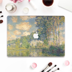 Può includere: Una cover per laptop con un dipinto di paesaggio con un fiume, alberi e un cielo nuvoloso. L'opera d'arte è nei toni del verde, blu e giallo. Il logo Apple è visibile. Circondato da pennelli per il trucco, una tazza di caffè e altri prodotti di bellezza.