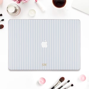May include: A light blue and white striped laptop case with the Apple logo. The case has the initials "SW" in gold. The case is surrounded by makeup brushes, a cup of coffee, and beauty products.