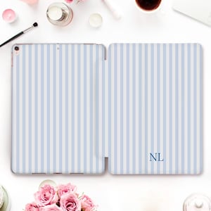 May include: An open iPad case with a light blue and white striped pattern. The initials "NL" are printed in a matching blue color on the right side of the case. The case is shown open, revealing the iPad screen.