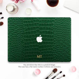 Può includere: Una custodia per laptop verde scuro con una stampa effetto pelle di coccodrillo e un logo Apple bianco al centro. Le lettere MT sono stampate in oro sotto il logo. La custodia ha una superficie opaca piatta priva di texture fisica.