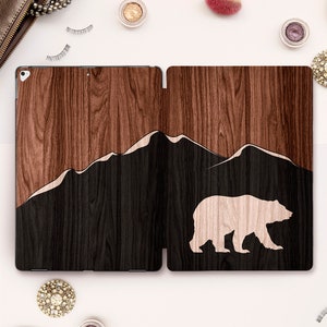 Puede incluir: Una funda para iPad de madera marrón con una silueta de montaña negra y una silueta de oso blanco. La funda está hecha de dos piezas que se unen a presión.