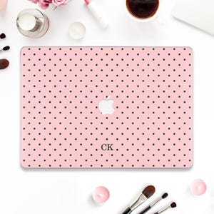 May include: A pink laptop case with a black polka dot pattern. The Apple logo is visible in white. The initials "CK" are printed in black below the logo. The case is surrounded by makeup and beauty products.