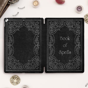 Peut inclure: Une housse pour iPad en cuir noir avec un motif en relief argenté. La housse est ouverte et la couverture avant porte l'inscription "Book of Spells".