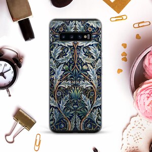 Peut inclure: Une coque de smartphone avec un motif floral dans les tons de bleu, vert et marron. Le motif présente des feuilles et des fleurs stylisées sur un fond bleu foncé. La coque est entourée d'autres objets, dont une horloge et des trombones.