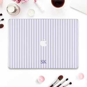 May include: A laptop case with a vertical striped pattern in light purple and white. The Apple logo is visible, along with the initials "SK" in a dark purple font. The case is surrounded by makeup and beauty products.