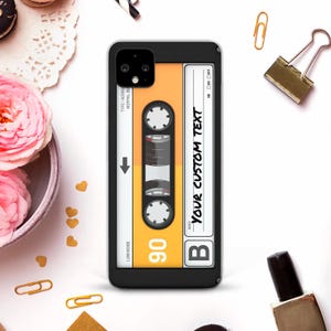 Pode incluir: Capa de telefone preta com um design de fita cassete retrô. A fita cassete tem um fundo laranja com detalhes em branco e preto e o texto "YOUR CUSTOM TEXT". A capa tem uma borda preta e um recorte para a câmera.