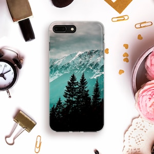 May include: A clear phone case with a scenic image of snow-capped mountains and a forest of dark green trees in the foreground. The sky is a gradient of blue and gray.