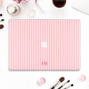 May include: Pink and white striped laptop case with the Apple logo and the initials "SM". The case is surrounded by makeup brushes, a cup of coffee, and beauty products. The case is a stylish accessory.