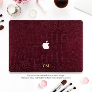May include: A burgundy laptop case with a faux crocodile skin texture. The Apple logo is white. The initials "GM" are at the bottom. The text reads: "Pay attention that this is a printed image. The case has a flat matte surface without any textures."