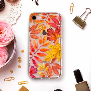 以下が含まれることがあります： 赤、オレンジ、黄色で描かれた秋の葉の水彩デザインが特徴の透明なiPhoneケース。「iPhone」の文字がケースに表示されています。文房具や装飾品に囲まれています。