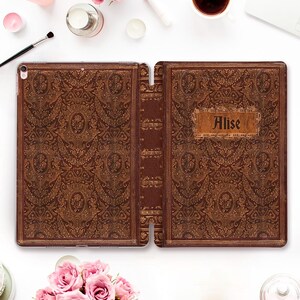 Puede incluir: Funda de iPad de cuero marrón con un diseño floral en relieve. La funda está abierta, revelando el interior y el lomo. El nombre "Alise" está impreso en una etiqueta rectangular en la portada. La funda tiene una estética de libro antiguo.