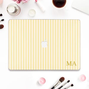 May include: A yellow and white striped laptop case with the Apple logo cutout. The initials "MA" are printed in gold on the lower right corner. Makeup brushes, a cup of coffee, and beauty products surround the laptop.